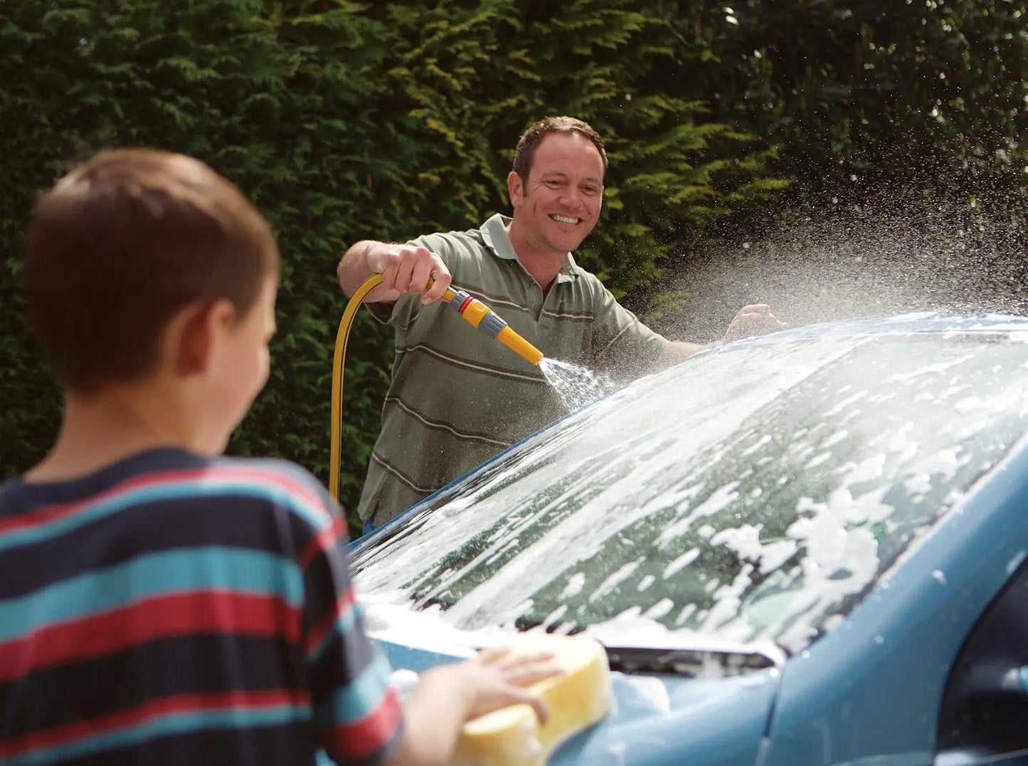 The Hozelock Hose Nozzle is an adjustable spray fitting designed for quick and efficient watering, ideal for a variety of garden tasks. Man and child washing a car together outdoors