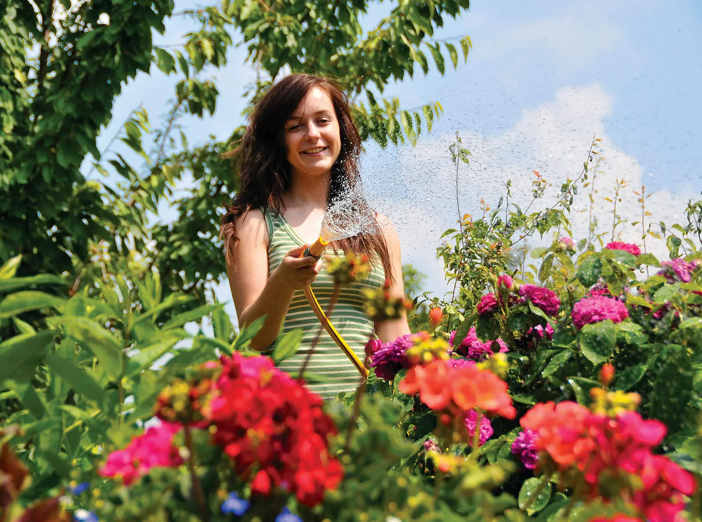 The Hozelock Hose Nozzle is an adjustable spray fitting designed for quick and efficient watering, ideal for a variety of garden tasks. Woman watering flowers in a garden with a blue sky and greenery in the background